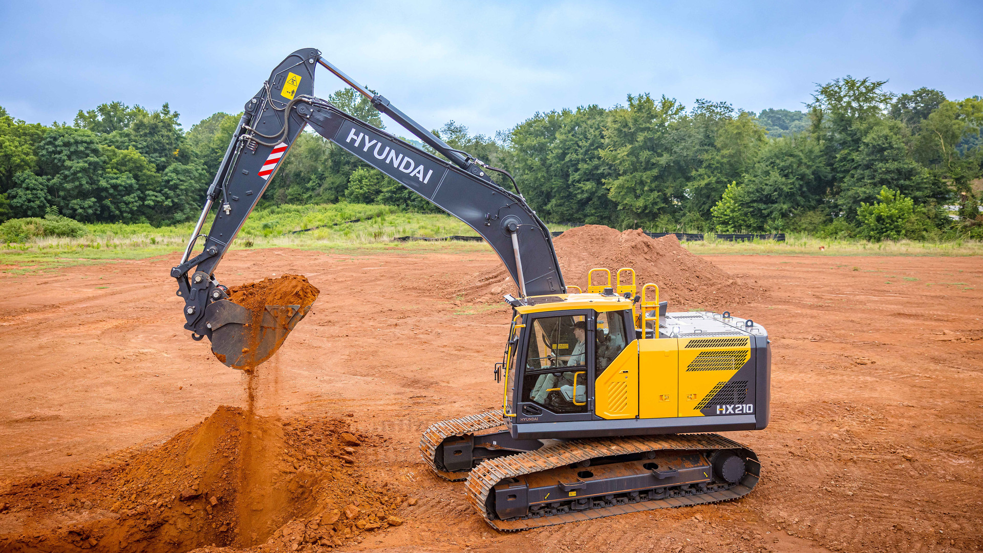 Hyundai HX210L excavator tipping a bucket of red soil on a cleared site, HX210 badge on the side of the upperstructure