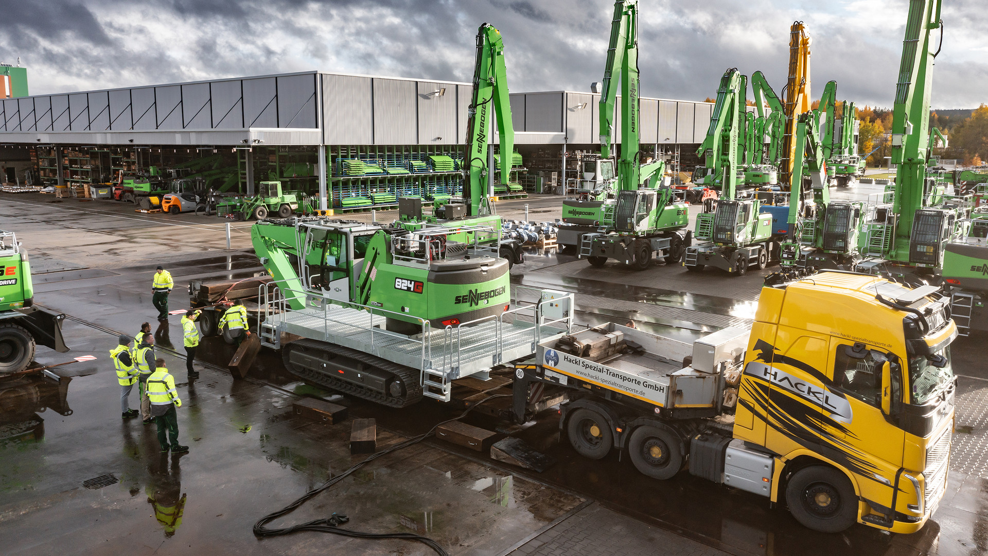 SENNEBOGEN 824 G Electro material handler loaded whole on a low-loader at the SENNEBOGEN plant, 824 G badge visible, with the full-length grated service walkways that take it out to five metres wide