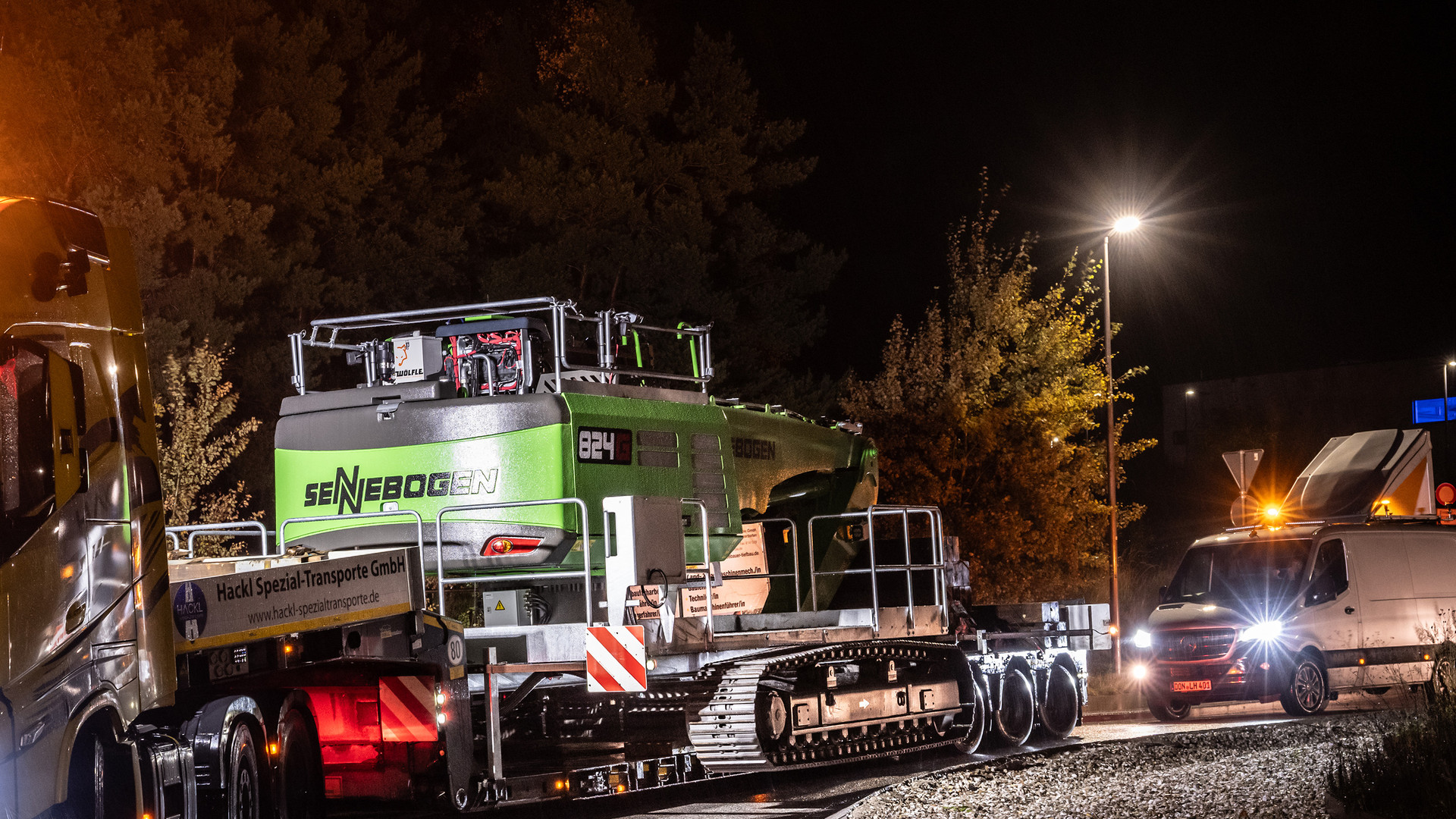 SENNEBOGEN 824 G Electro on a Hackl low-loader during the night leg of the move, the 824 G badge lit and an escort vehicle following on the roundabout